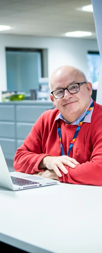 Smiling staff member in a red jumper sitting at a desk with a laptop in a modern office setting at Glasgow Kelvin College Smiling staff member in a red jumper sitting at a desk with a laptop in a modern office setting at Glasgow Kelvin College
